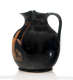 Black vessel with a round body and flared lip connected by a slim handle. The glazed surface is extensively cracked, with a barely visible orange illustration to the left.