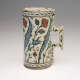 A rounded cream colored vessel with a square handle, decorated with geometric and flower designs in blue, green, and red.