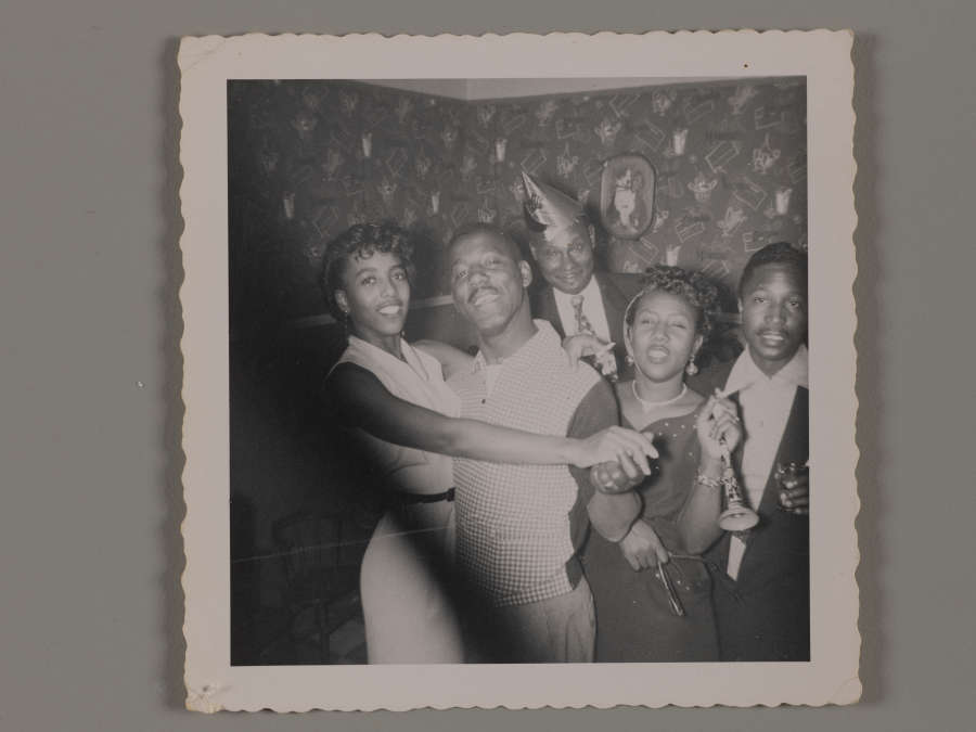 Black and white party snapshot of five smiling dark-skinned, well-dressed adults. One man wears a pointed paper hat, the couple at left are dancing, and another woman holds a noisemaker.