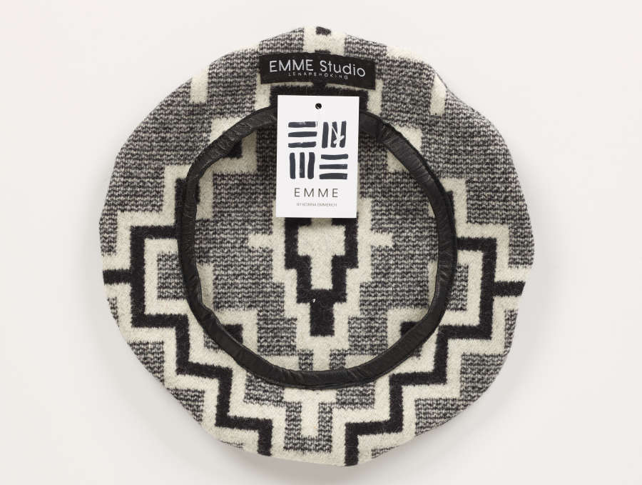 Underside of a gray tweed beret with geometric patterning and black leather lining the hat’s opening. An attached white rectangular brand tag below the brand label reads 'EMME’ with line motifs.  
