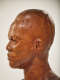 Close-up profile view of a textured, but glossy, wooden-carved bust depicting a male figure with afrocentric facial features, he has a stern expression and an intense gaze.