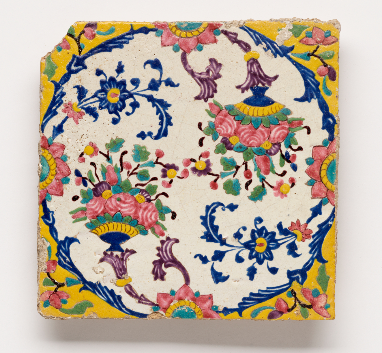 Square wall tile with a chipped upper left corner. It features floral pink, green, and yellow motifs connecting into swirling blue motifs to form a circle. The corners are yellow.