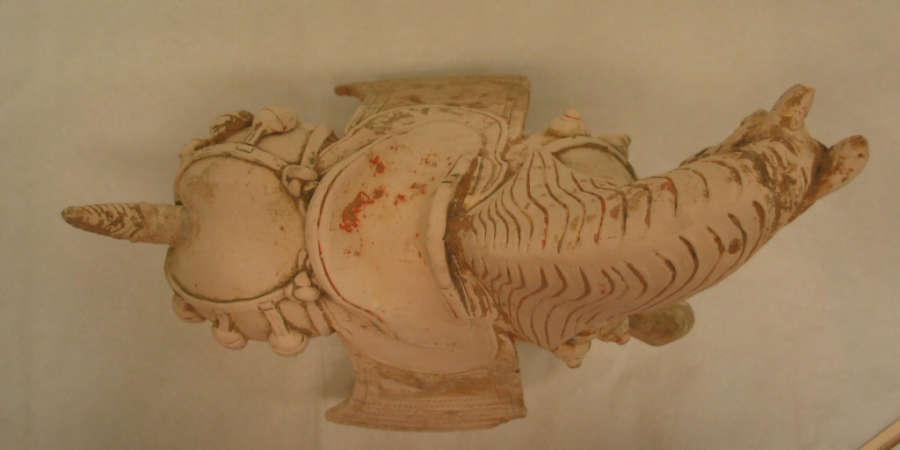 Top-view of a white ceramic sculpture of a horse, looking aside, with its mane flowing down its neck, a layered saddle, a wound tail, and ornamental belts.