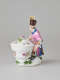 A sculptural figure kneeling next to a basket. Figure wears bright, elaborate clothing with a hat with a feather. There are floral decorations on the figure’s clothing and basket.