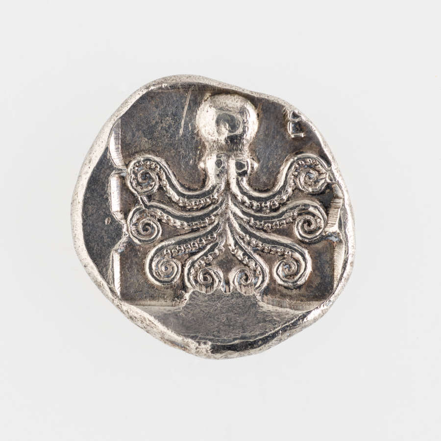 A round silver coin with a raised imprint of an octopus with curling tentacles. Although the octopus is symmetrical, it is placed off-center on the coin.