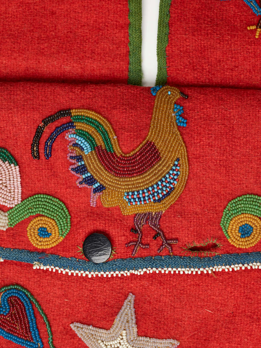 Detail of the red bag's body, showing an intricately beaded chicken as well as a small button and a cream lined embellishment sit in between the blue rimmed bag opening.