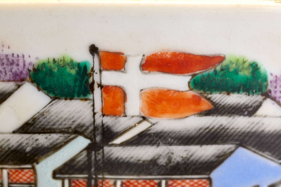 A close up of a painted ceramic with a red and white cross flag on a pole, set against a background of black rooftops and green foliage.