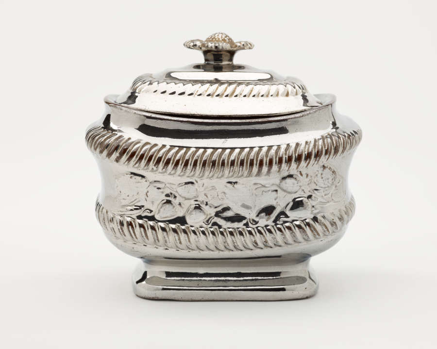 A silver sugar bowl with a decorative lid and a rounded square body with sculptural decorations.