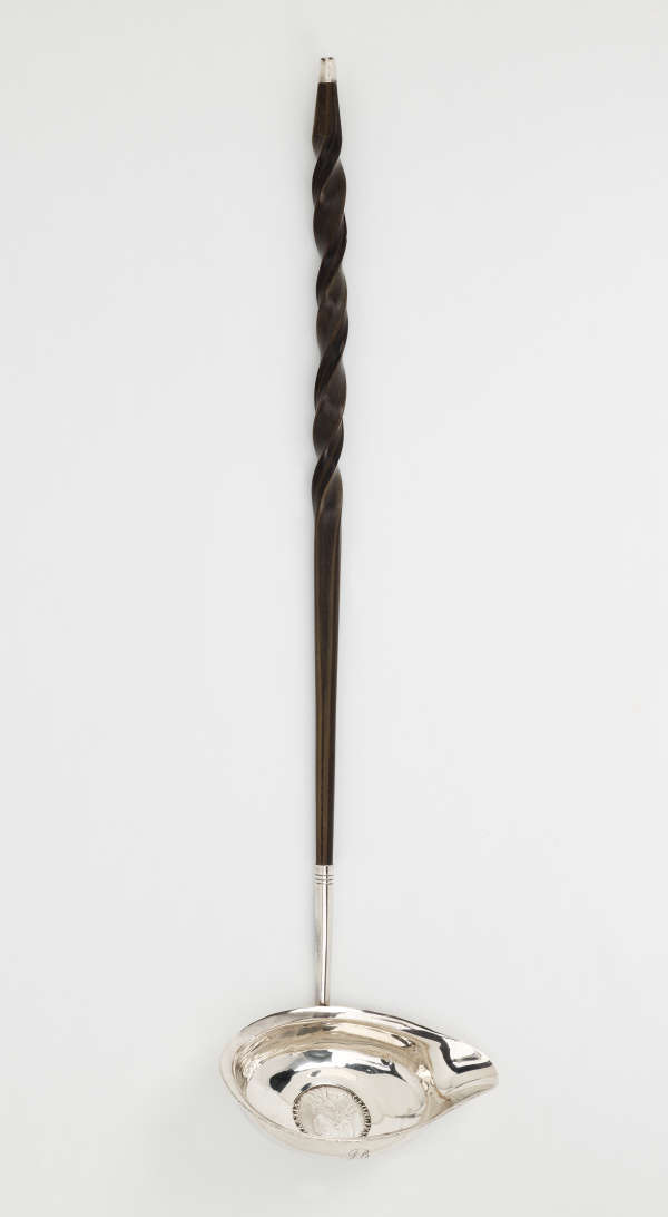 A silver and baleen coin ladle. The handle is twisted and dark and the ladle is silver with a small coin in the center.