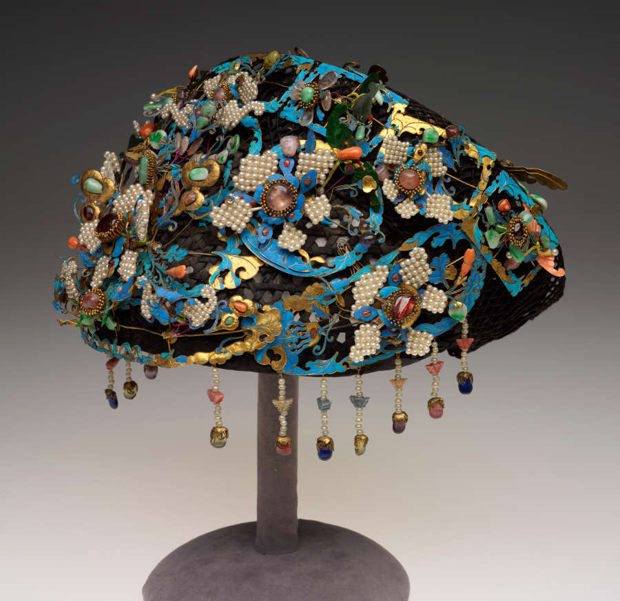 Back view of the headdress on a gray stand. The headdress features blue and gold leaf frames, pearlescent beaded stone-set flowers, flower and insect shaped stones and hanging beaded ornaments.
