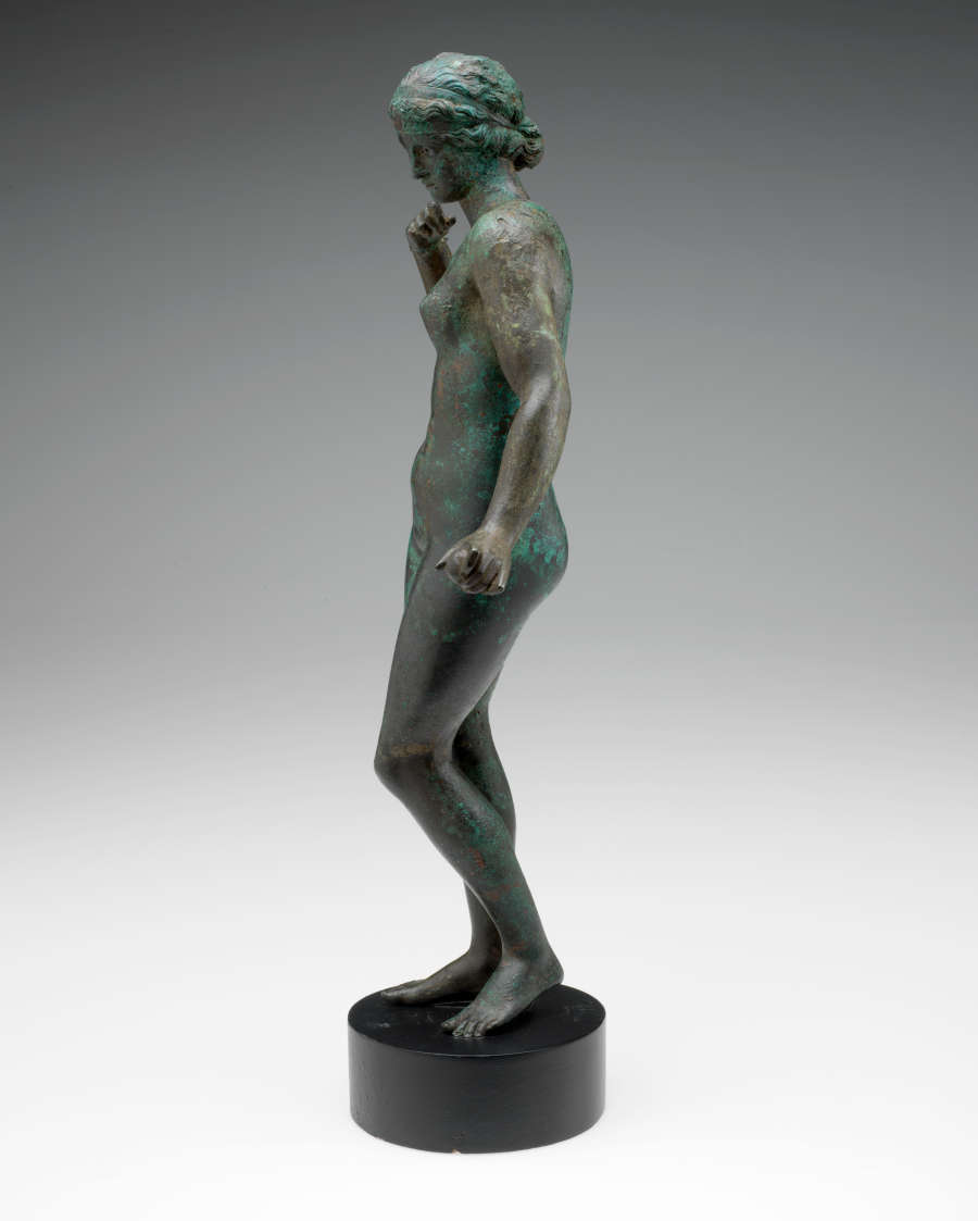 Side view of a tarnished bronze statue of a standing crowned nude woman with one arm bent towards her shoulder and the other held out, standing on a circular pedestal.