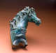 Greenish-gray metal ornament of a horse’s head, turned, with curls of hair falling along the ridge of its neck and its saddle. The horse’s mouth is partially open.
