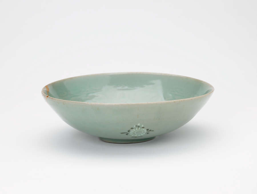 Side-view of a tilted, light-teal, stout glazed bowl, featuring green-blue floral motifs on the exterior, and a faded floral pattern on the interior, along with a narrow foot.