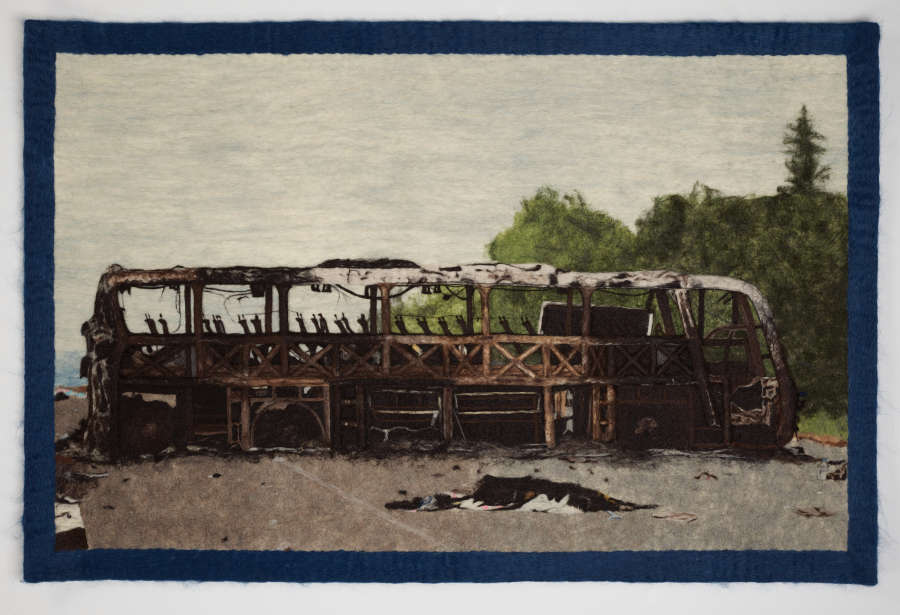  Textile depicting the charred remains of a bus with skeletal framework exposed. On the left most side of the background there are lush trees against an overcast sky.
