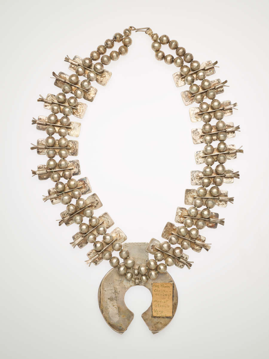 The back of a silver pendant necklace with two rows of silver beads throughout. On the central larger pendant there is a rectangular cream cloth on the right side of the semi-circle pendant.
