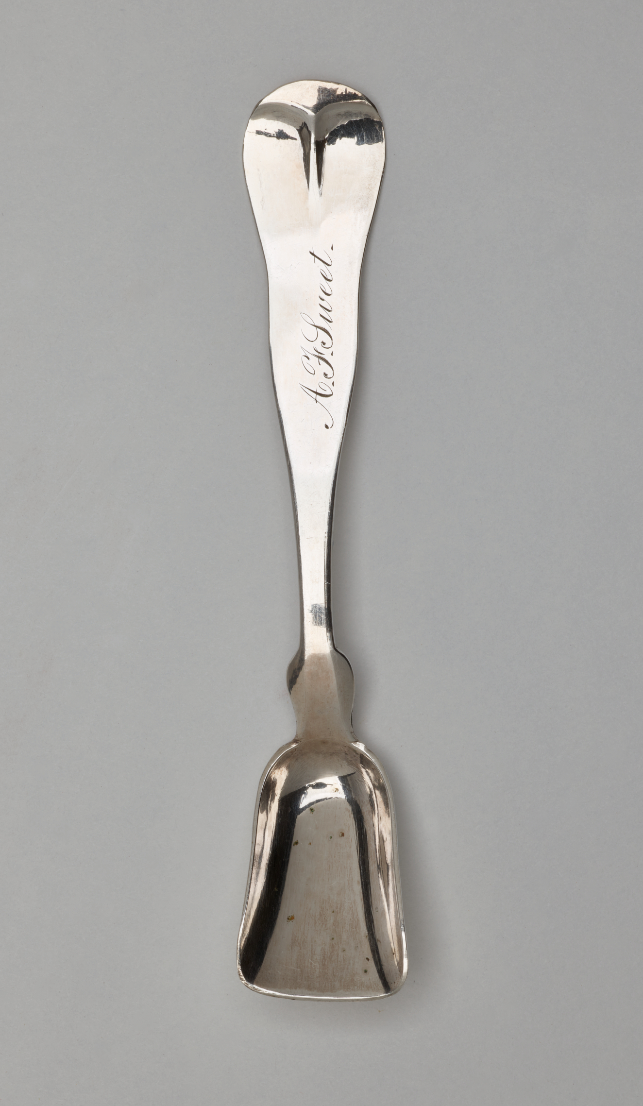 A silver salt spoon with a bowl that comes to a blunt flat tip and a neck that starts out thinner and goes to a thicker rounded handle.
