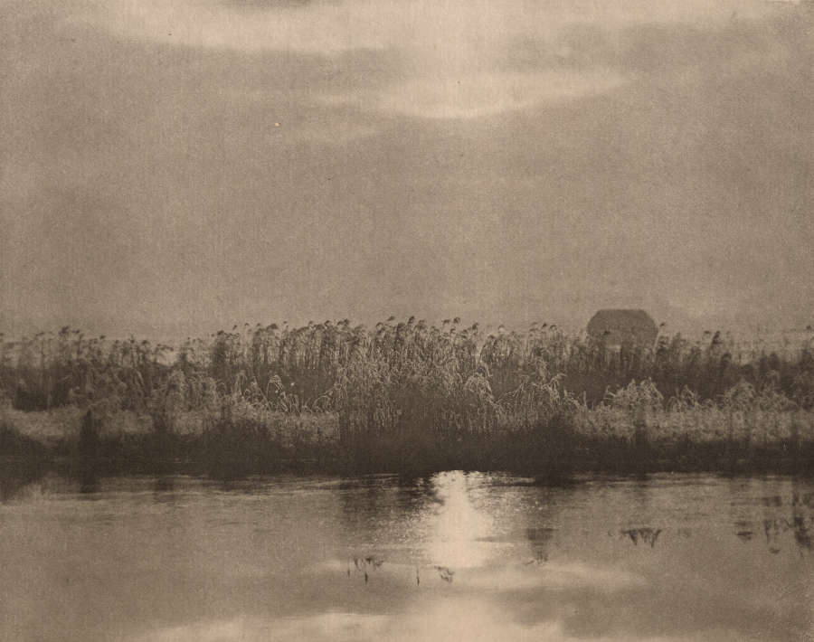 A black and white landscape with a tranquil body of water reflecting a grassy shoreline in the foreground and a small house set against a pale sky in the background.