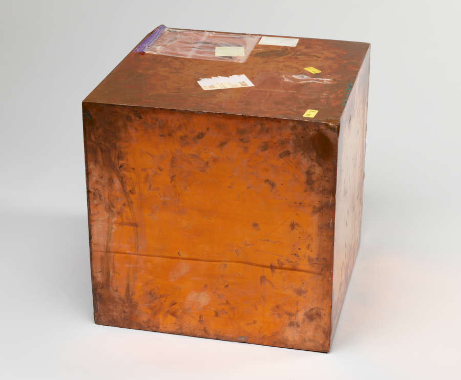 Image of Walead Beshty’s work: Copper box covered in various markings and shipping label stickers. The box is discolored in some areas.