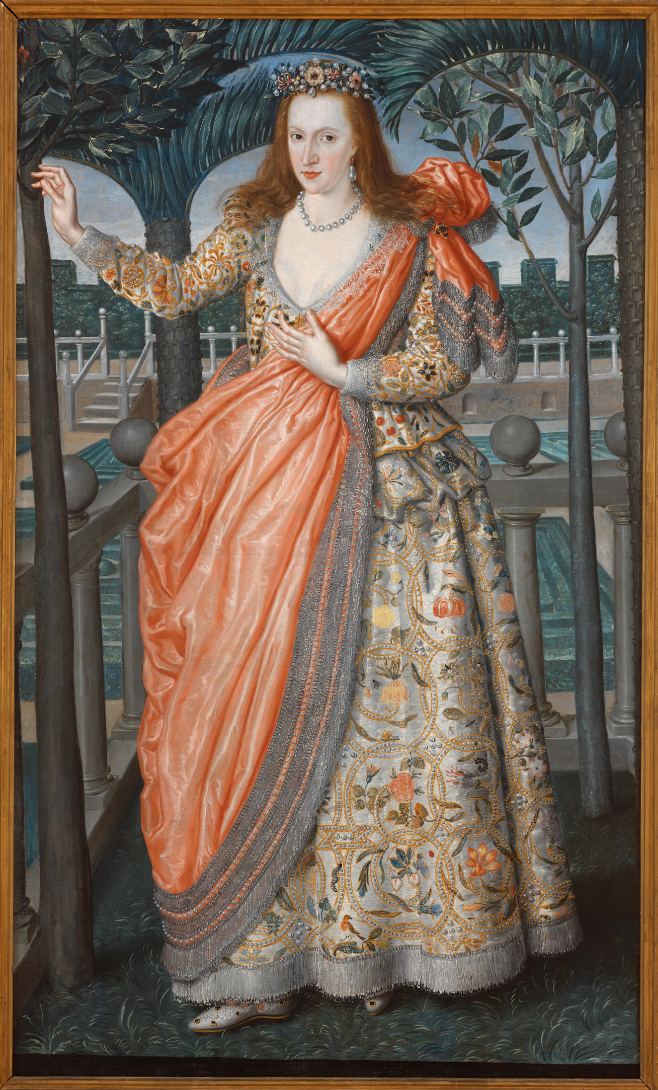 A woman in an opulent embroidered gown with florals and gold detailing and draped in a flowing orange shawl, posing elegantly in a garden. 
