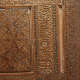 A close up view of a carved wooden panel with intricate calligraphic inscriptions in a center column, surrounded by geometric, floral borders. 