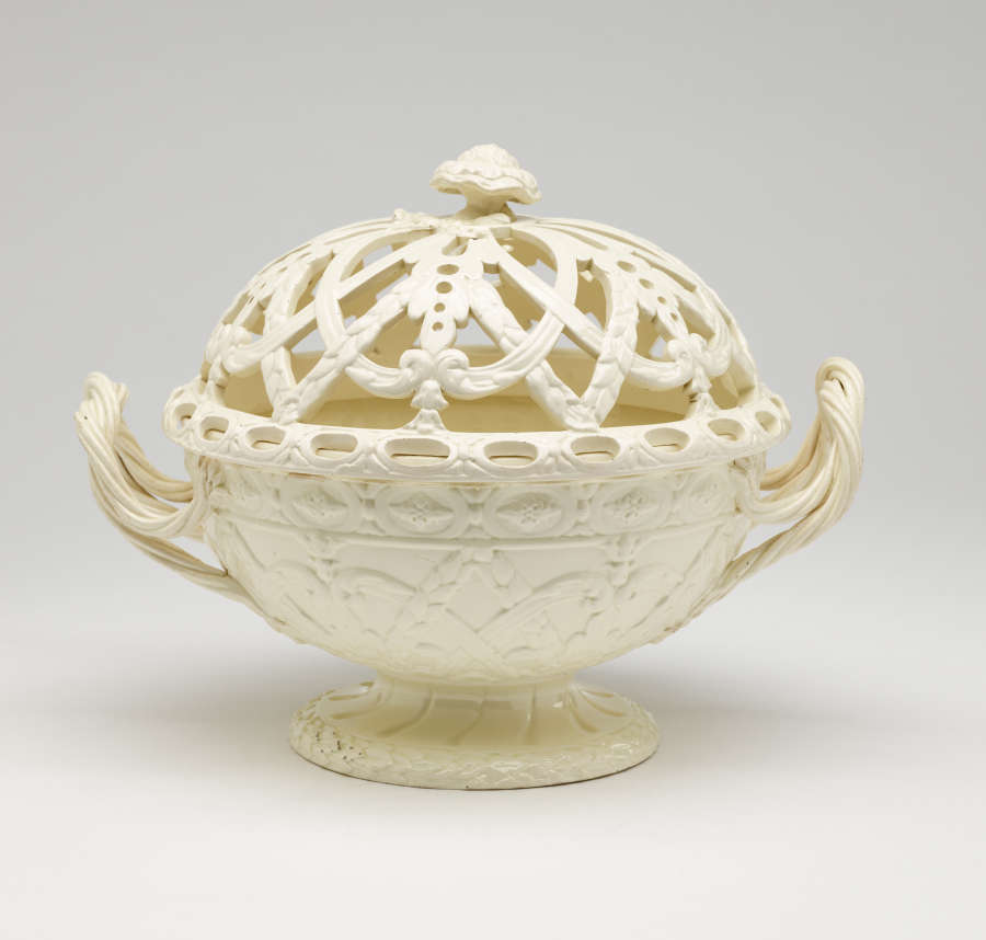 A cream-colored basket with sculptural woven handles, sculpted foot, and a lid with cutout designs.