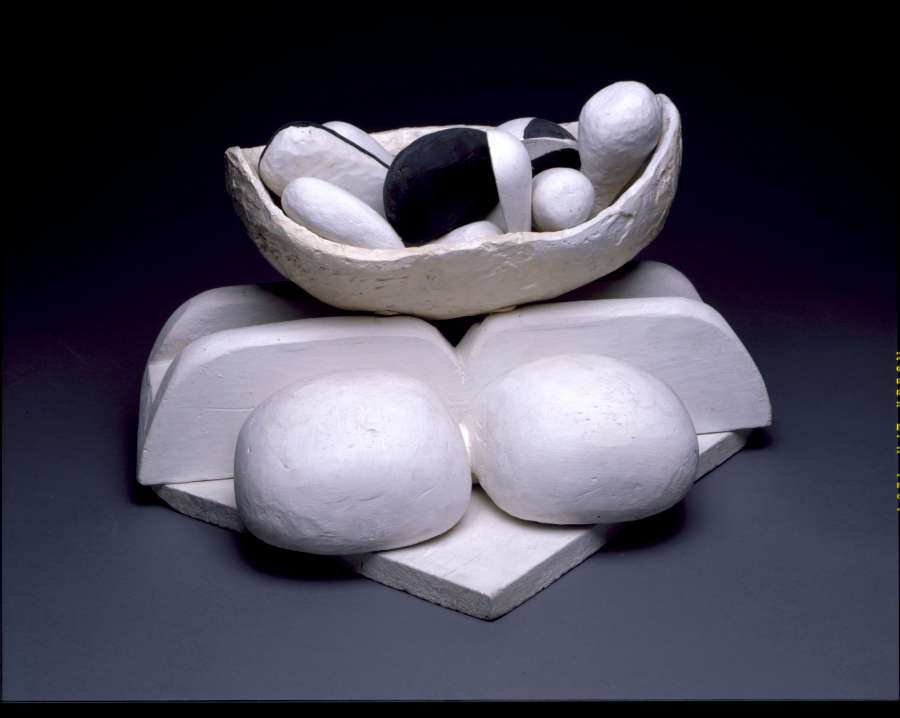 Against a dark background, a white bowl, containing several smaller orbs, balances on top of a square platform with two channeled wedges, flanked by two larger white round objects. 
