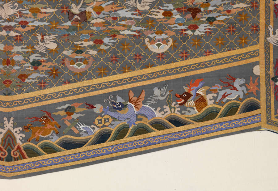 Right border detail of the triangular tail of the gray-blue robes back featuring illustrations of wavy landscape, dragons, birds, and animals encased by blue and gold patterned stripes