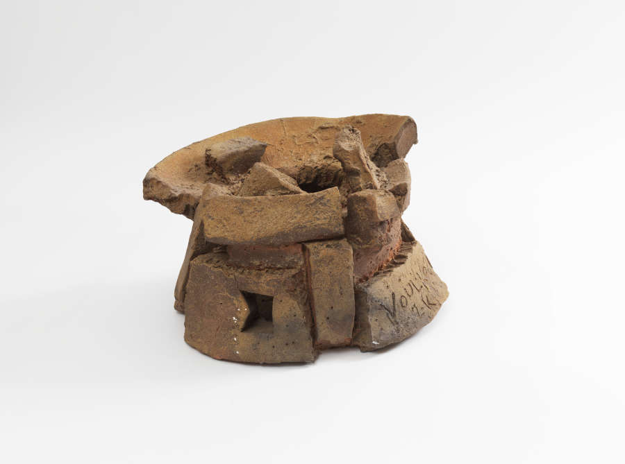 Abstract brown ceramic sculptural form composed of randomly stacked slabs resembling bricks with rectangular openings, deep cracks, and the artist’s surname inscribed on one slab.