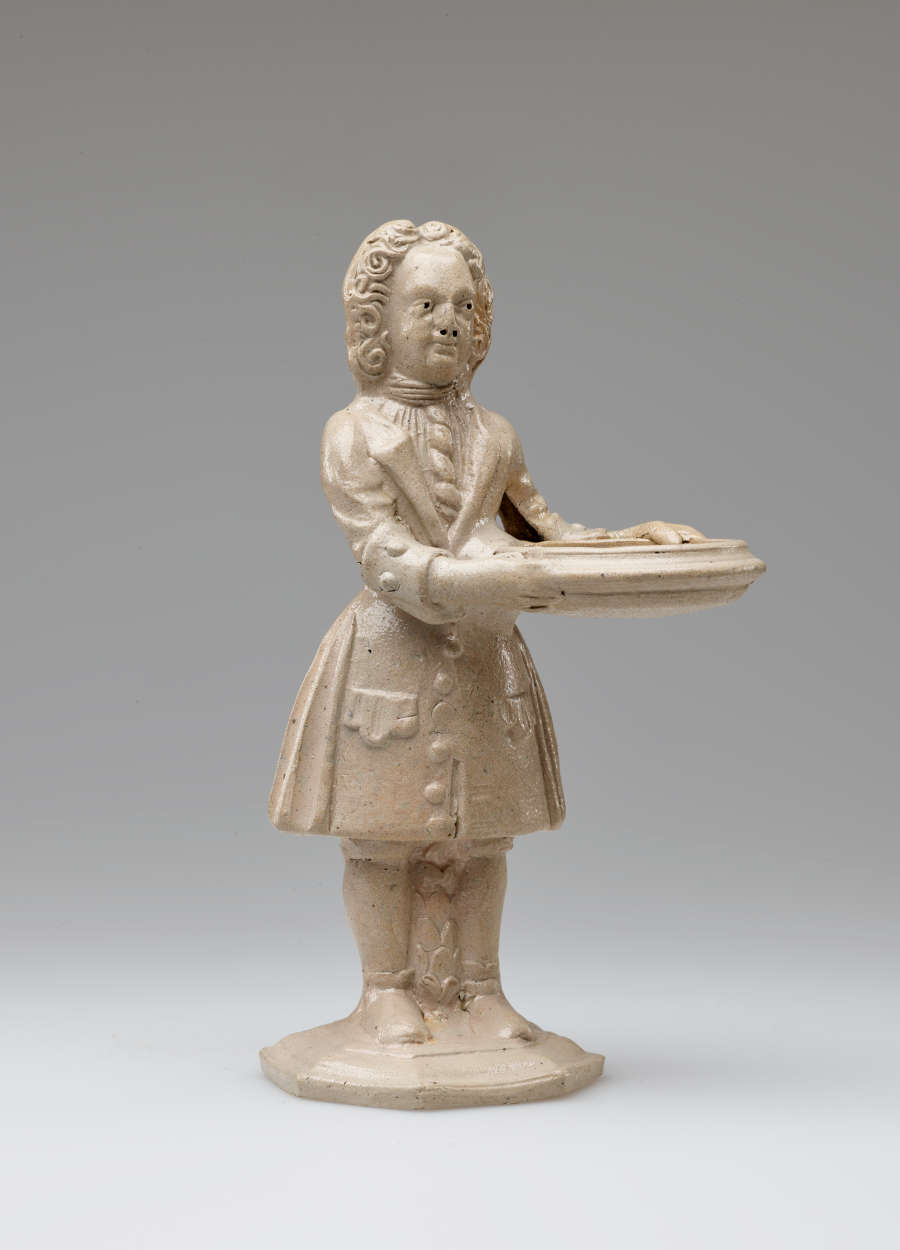 A cream colored sculptural figure holding a vessel, dressed in historical clothing with tightly curled hair.
