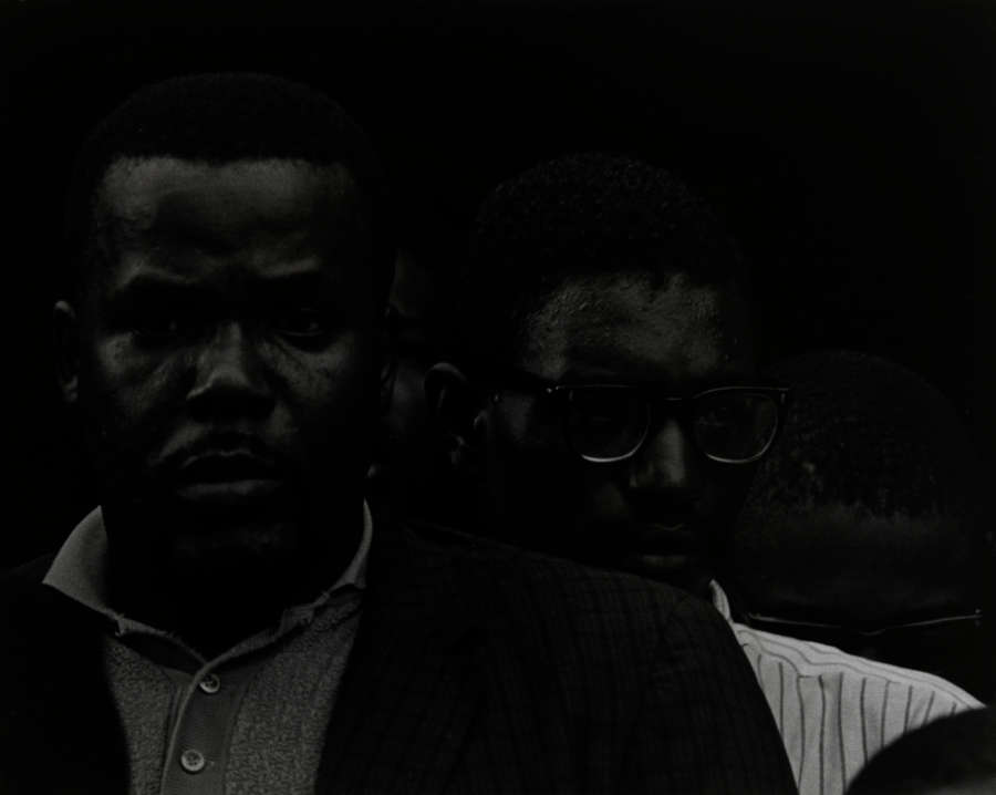 A dark, low contrast black-and-white photograph capturing two nearly imperceptible dark-skinned men gazing at the viewer with inscrutable expressions. One is wearing glasses and partially obscured behind the other. 