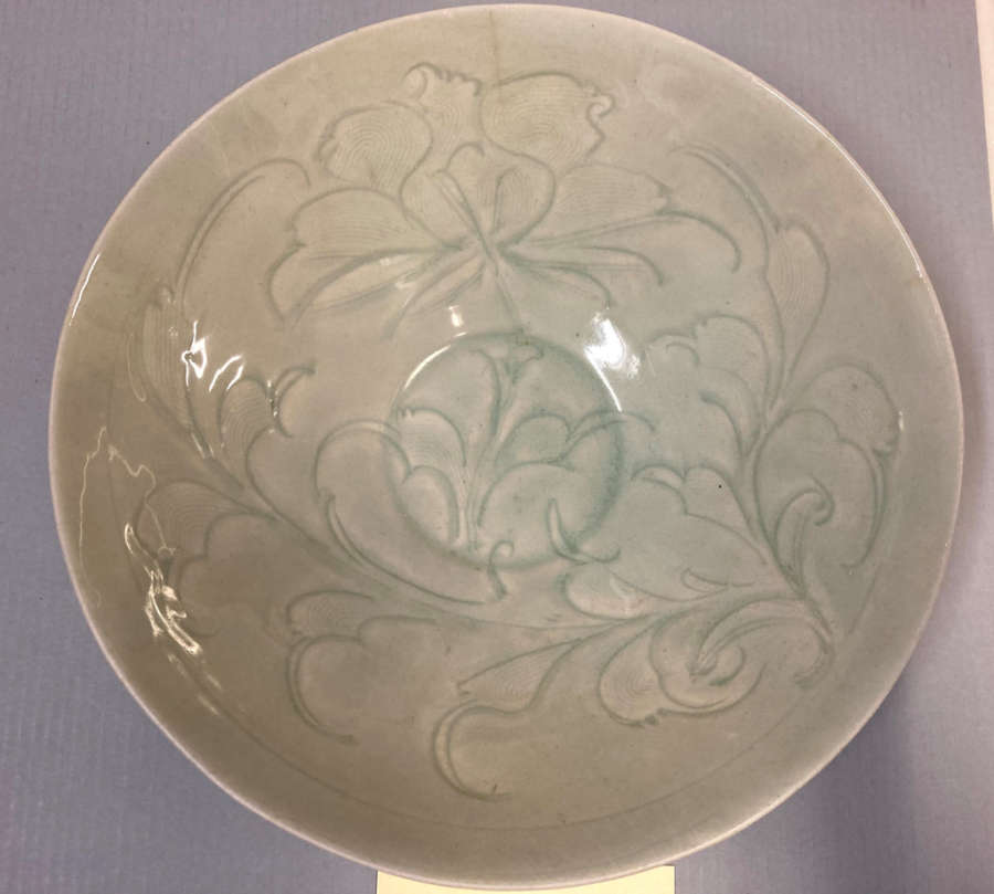 Top view of a round glossy pale gray-green bowl with carved floral motifs which flow into one another. Its base is circular. The plate sits on a purple-gray background.