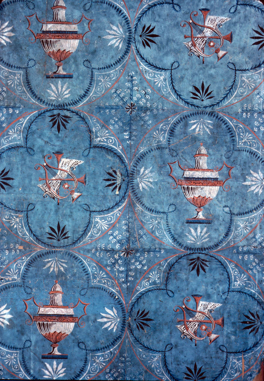 Antique wallpaper made of individual teal tiles featuring urns and musical instruments surrounded by foliage motifs within a rounded four-leaf clover. The instrument tiles and urn tiles are arranged alternatingly.