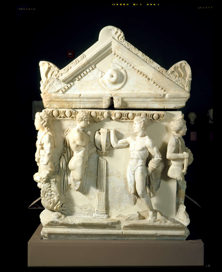 A panel of a marble coffin with a triangular roof and deep set carvings of standing figures engaging in conversation. 