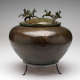 A brown-green patinated metal bowl with decorative engravings on its onion-shaped body, sitting on a three-legged stand. Metal-cast winged horses dance along the rim of the bowl’s domed lid. 