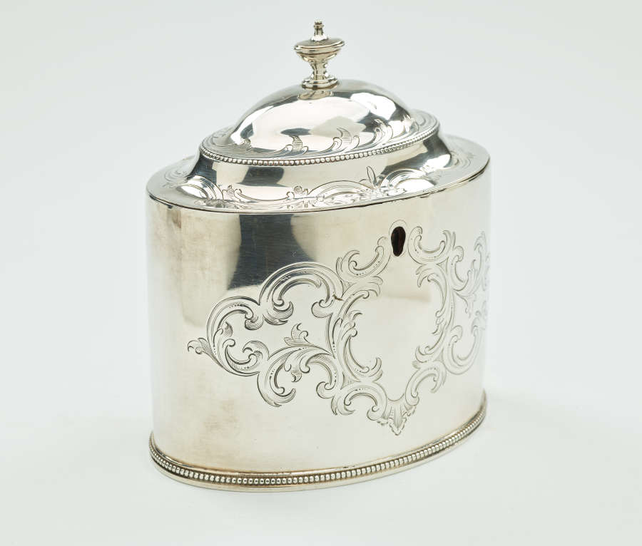 A silver tea caddy with a lid and sculptural finial. There are swirling engravings along the body and lid.