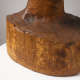 Back view of a carved wooden bust depicting the head of a male figure. The wood is cracked while retaining shine, the sculpture also features carving marks throughout.