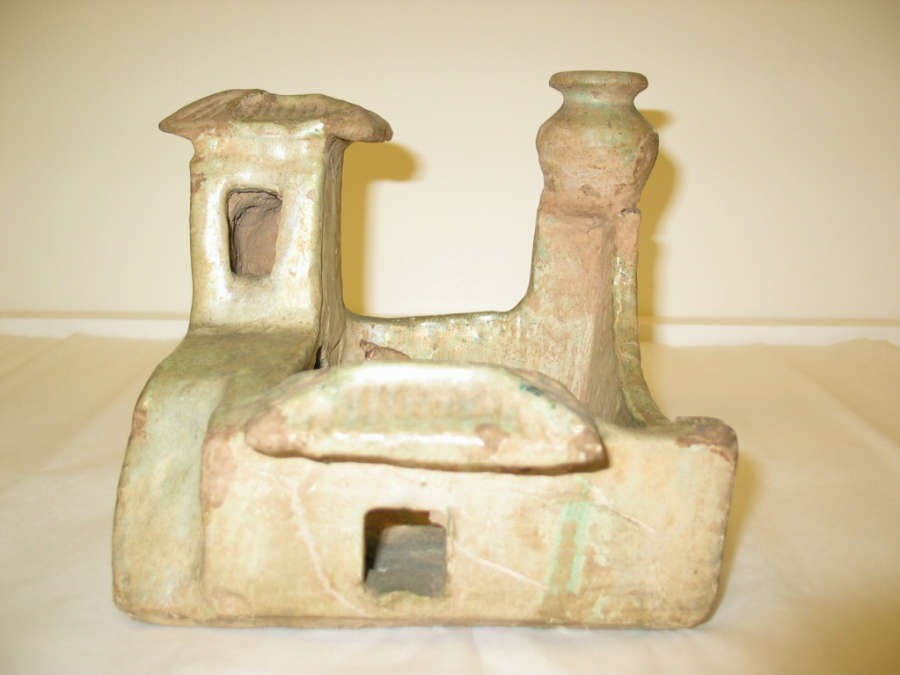 Front-view of a pale-green ceramic sculpture in storage of a farmyard with a courtyard, roofed entrance and two towers, one with a room, and another with a pot upon it.