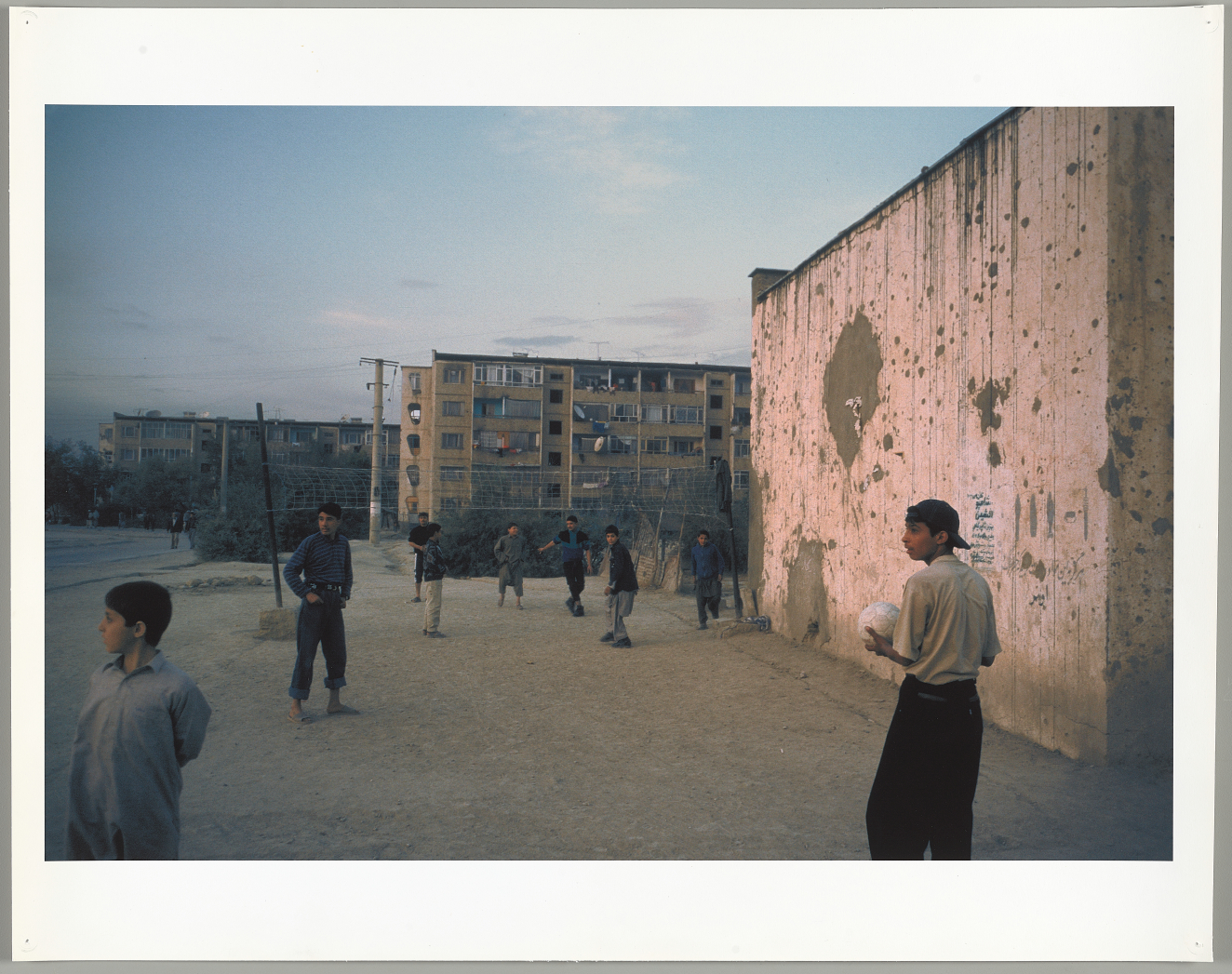 A group of young teenagers play ball near a pink bullet-scarred wall dominating the right side, with a damaged apartment in the background, set against a dusky blue sky.