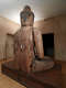 An angled rear view of a large wooden Buddha statue, seated on a wooden platform in a warmly lit room with visible signs of cracks and repairs.