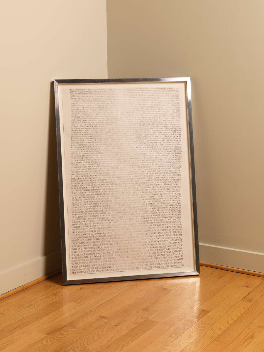 Image of Fiona Banner’s work (the side with the text in graphite): Framed work sits on the floor, leaning against a corner. The front of the work is visible and features a block of small text in graphite. The frame is silver.