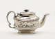 A silver teapot with a decorative handle, a rounded square body and spout with sculptural decorations.