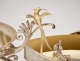 Detail of golden bird with long two-part tail and long, thin curved beak perched on an ornate silver decorative scroll attached to a shallow dish above a winged cherub figure. 