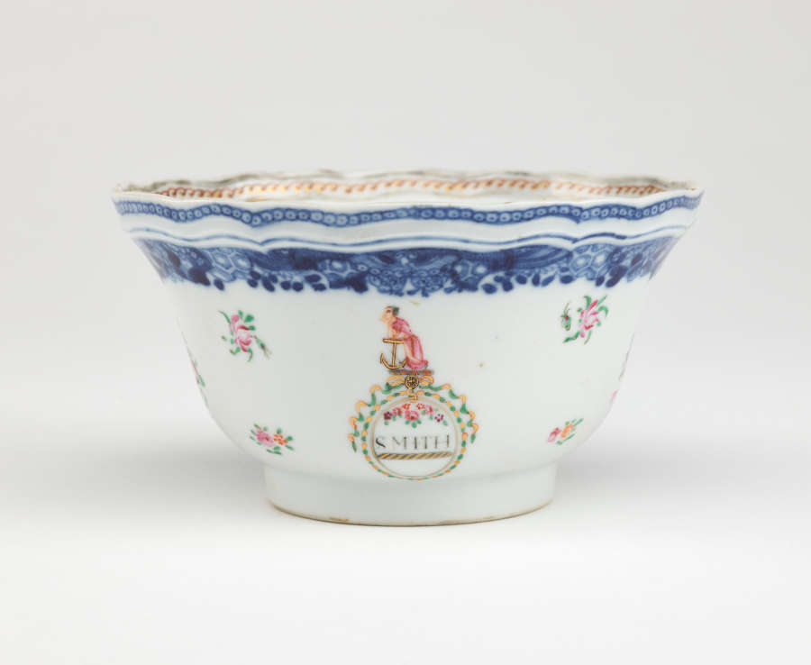 A bowl with glided, blue, green, and pink decorations. The body is decorated with flowers and a figure resting on an anchor. Word below the figure says “SMITH.”