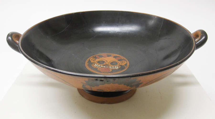 Partial top-view of a cup with a wide mouth showing two handles peeking out from the edges. Its interior is glossy black with a terracotta illustration of an animal face. 