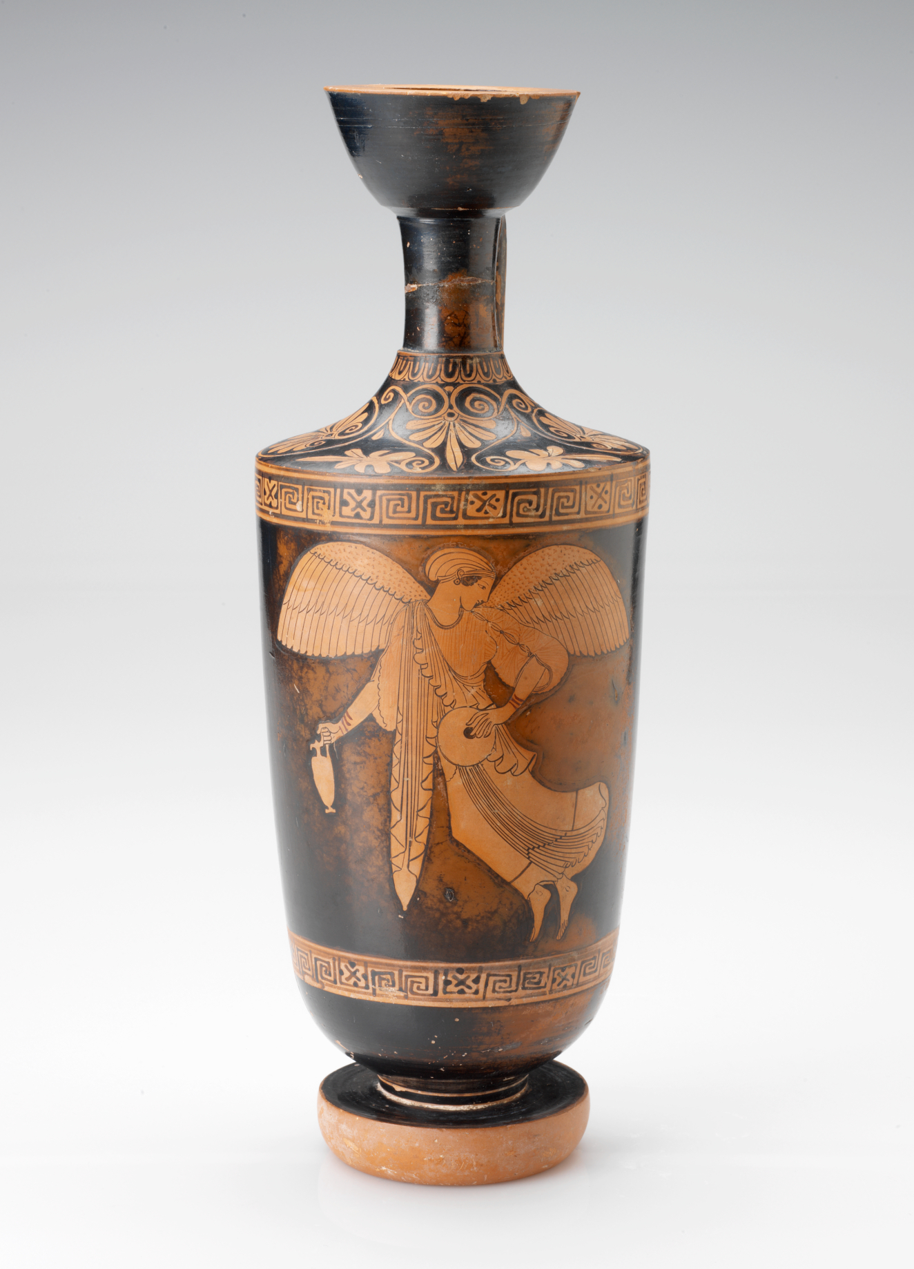 Tall black vessel with a narrow neck and wide mouth, whose surface features orange floral patterns above a winged figure holding a vessel and flask framed by geometric borders.