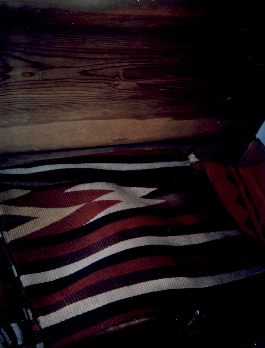 Vintage photograph showing a corner view of two blankets, a patterned red, blue, and beige blanket and a red woven blanket with green triangular designs, inside an open wooden box. 
