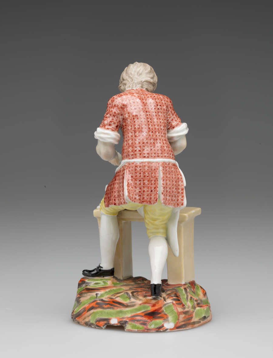 A sculpture of a figure in historical clothing and a table, a sack, and broken egg shells. The ground is brightly colored red, brown, and green.