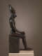 The side view of a dark statue of a human figure seated on a throne, with a falcon head and tall conical crown.