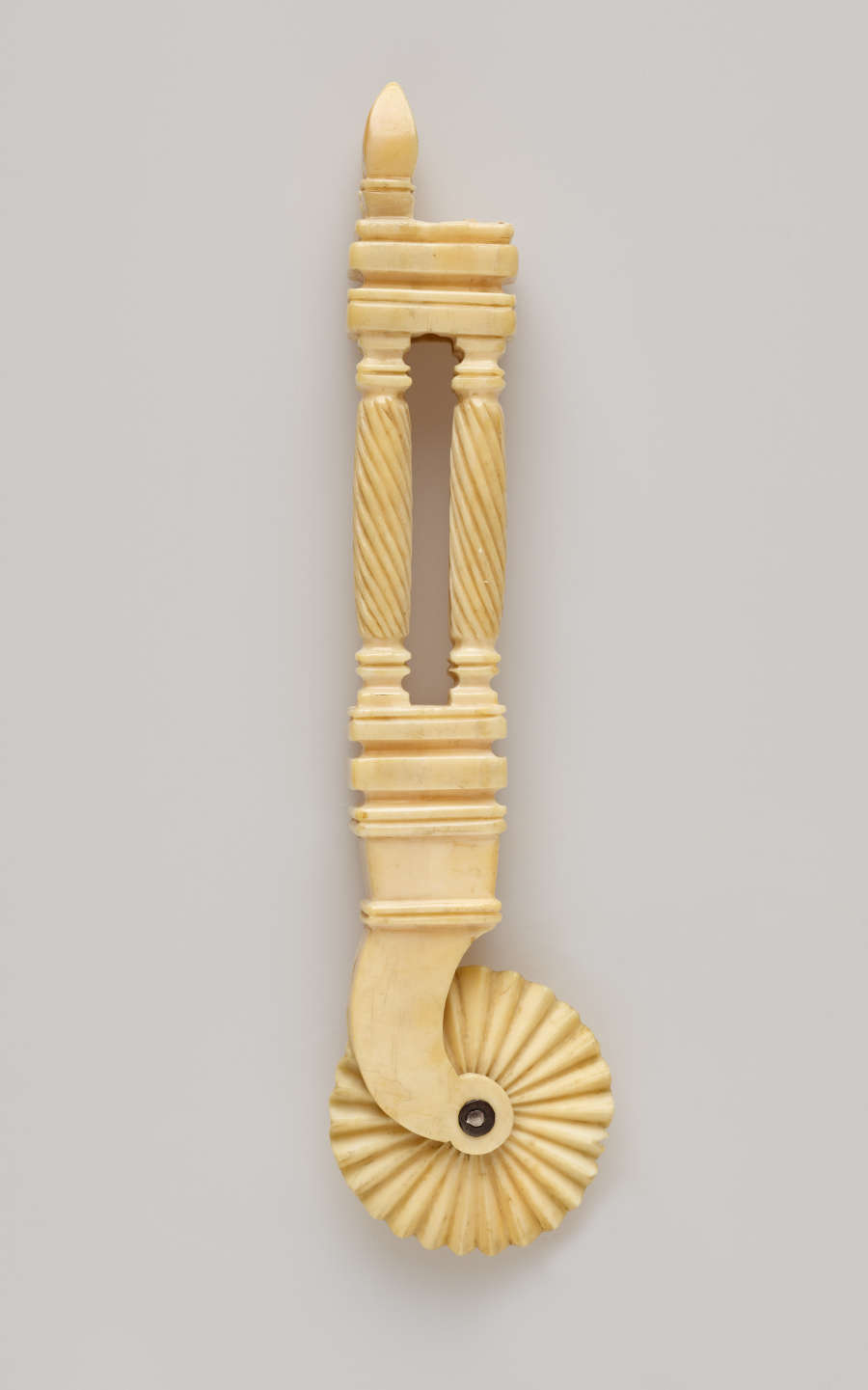 A tan colored tool with decorative handle and a textured wheel. The handle has a cutout in the middle of it.