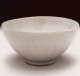 Side view of a glossy white ceramic bowl with a small round foot and overlapping lotus petal pattern engraved on its sides. Its mouth is round but uneven.